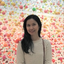 Thsi image shows Shirleyana standing in front of a pastel pink and yellow backdrop. She is wearing a white jumper and smiling at the camera.