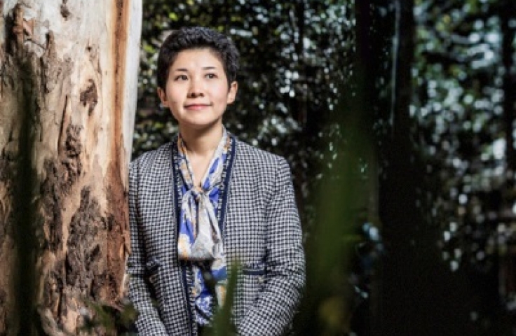 Xiaoqi leans against a gum tree. She is dressed in a black and white check blazer and is looking up from the camera.