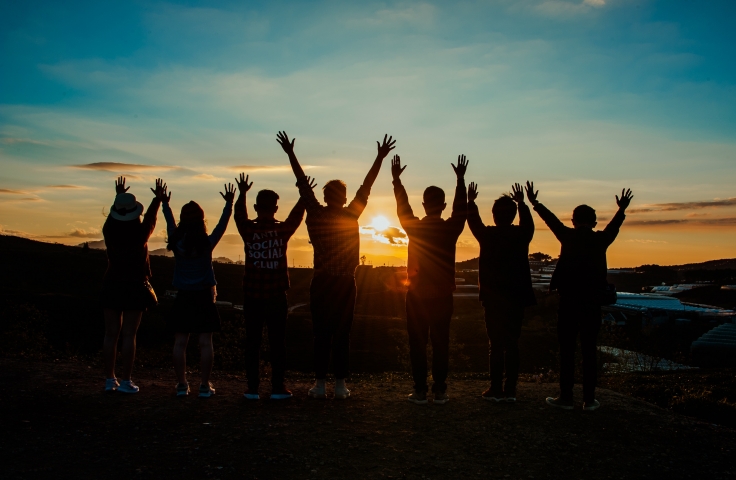 Seven people are standing in a line. They are facing the sunset with both arms up in the air.