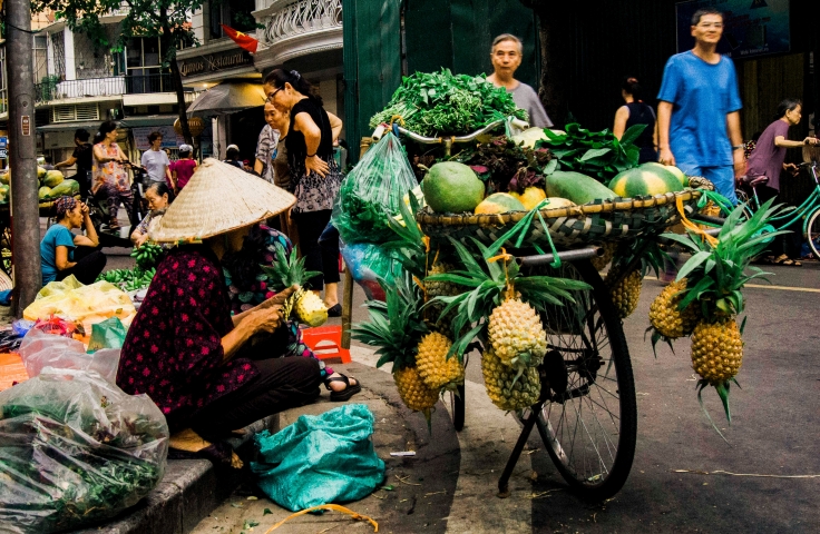A street vendor is sitting next to a road with a cart of vegetables for sale. 