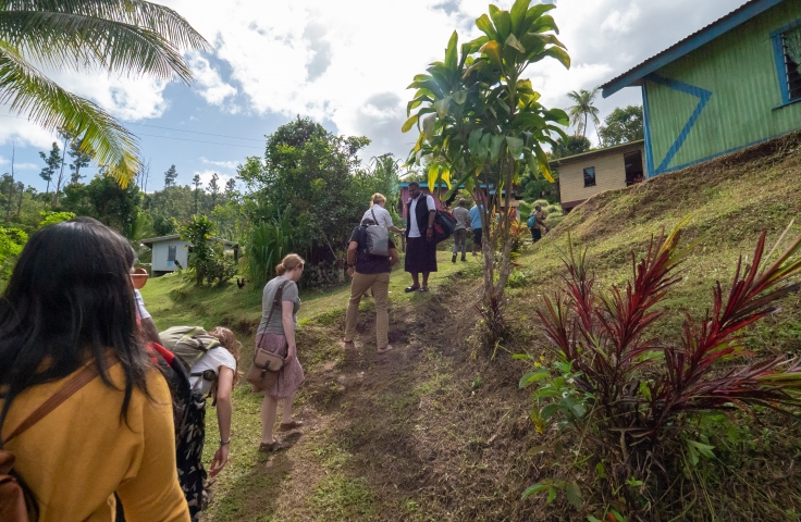 In the foreground a group can be seen walking away from the camera. In the background are houses located in a village.