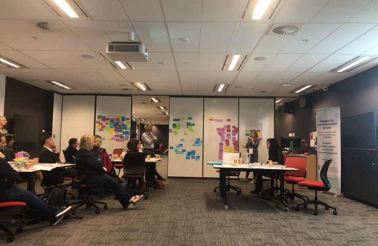 A profile view of a classroom. Participants are facing the front. Two facilitators are leading discussion. A wall is covered in small sticky notes.  