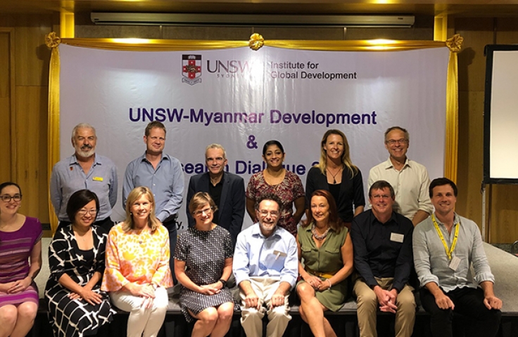 13 UNSW staff are seated on a stage facing the camera. They are seated in front of a banner that reads "UNSW Myanmar Development"