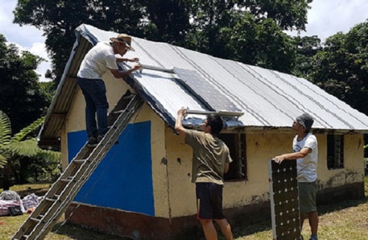 Three men are positioning solar panels on the roof of a small house. One man is on a ladder.