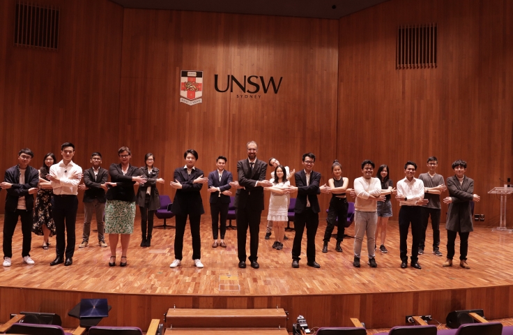 A group of participants stand on stage. They have their arms crossed as if holding hands with eachother. In the background, the UNSW logo is displayed on the wall.