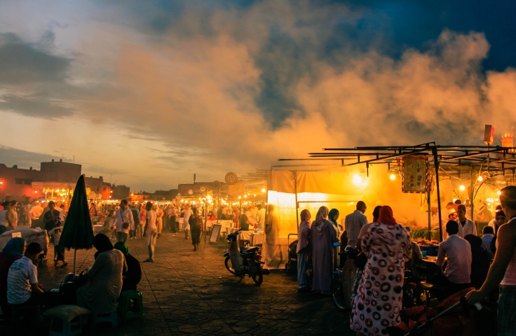 A landscape depicts a series of informal market stalls at sunset. There is smoke from a large cooking fire. People are milling around the stalls.