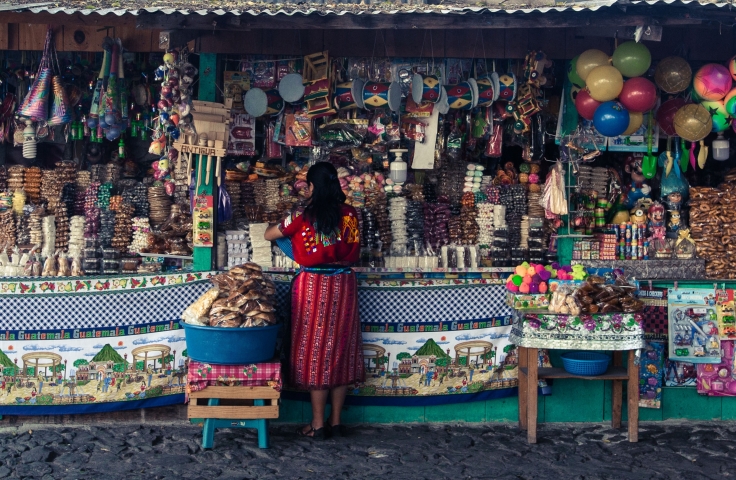 A woman stands with her back to the camera in front of a market stall in Guatemala. The stall is selling a variety of items.