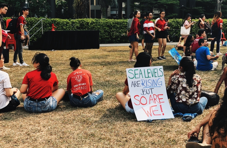 Protesters sit on the grass with a sign that reads "Sea Levels are Rising but So Are We"